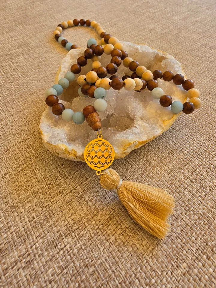 Le mala posé sur une géode, ses perles de bois clair et brun alternées d'amazonite dépoli, terminé par une breloque fleur de vie dorée et un pompon beige.