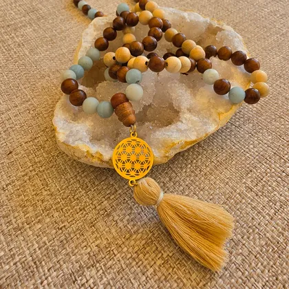 Le mala posé sur une géode, ses perles de bois clair et brun alternées d'amazonite dépoli, terminé par une breloque fleur de vie dorée et un pompon beige.