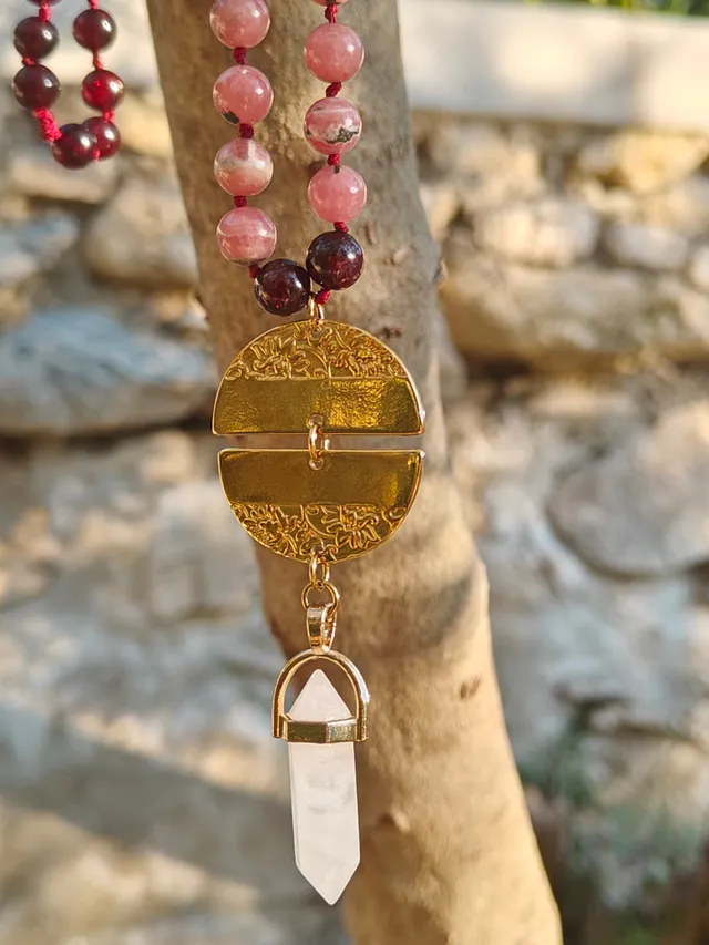 Gros plan d'un mala aux perles de rhodonite rose et de grenat, suspendu à une branche : un médaillon doré gravé de motifs floraux et une pointe de quartz clair terminent le collier